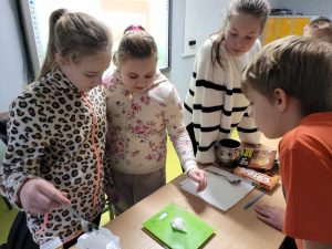 Uczniowie klasy czwartej stoją przy stole, na którym leży waga wraz z woreczkiem z cukrem. Dzieci przyglądają się wynikom pomiaru wagi.