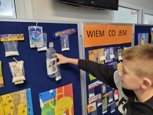 Uczeń przyczepia do tablicy butelkę po napoju "oshee" wraz z woreczkiem z cukrem. Obok przyczepione są inne produkty również z woreczkami.