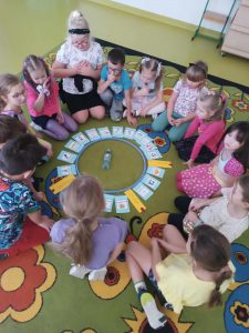 Uczniowie klasy pierwszej siedzą na podłodze w kręgu. Po środku leżą ułożone również po okręgu karteczki z różnymi napisami a wewnątrz tego kręgu leży butelka.