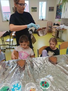 Dwie uczennice klasy pierwszej siedzą przy stole zakrytym folią aluminiową. Przed dziewczynkami na stole leżą foremki w kształcie choinek wypełnione czekoladą a dalej kubeczki z kolorowymi ozdobami do czekoladek. Za uczennicami stoi Pani trzymająca garnek z czekoladą.