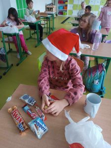 Uczennica siedzi w czapeczce mikołajkowej przy stoliku. Przed nią leżą słodkie upominki a obok stoi kubek otrzymany w prezencie.