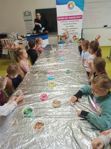 Uczniowie klasy pierwszej siedzą przy długim stole zakrytym folią aluminiową. Na stole ustawione są kubeczki z kolorowymi ozdobami do czekolady. W tle widać stojącą za kontuarem Panią w rękawiczkach. Obok niej stoi baner z napisem "MOBILNE WARSZTATY".