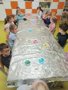 Uczniowie klasy pierwszej siedzą przy długim stole zakrytym folią aluminiową. Na stole ustawione są kubeczki z kolorowymi ozdobami do czekolady.