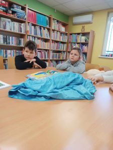 Dwoje uczniów klasy pierwszej siedzi za dużym stołem w gminnej bibliotece w Ostaszewie. Na stole leży niebieski materiał oraz otwarta książka.