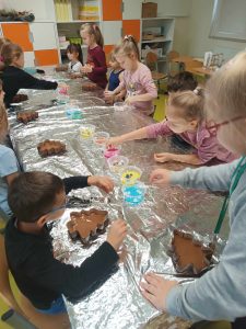 Uczniowie klasy pierwszej siedzą za długim stołem zakrytym folią aluminiową. Przed dziećmi ustawione są foremki w kształcie choinek wypełnione czekoladą. Dzieci ozdabiają swoje czekoladki kolorowymi posypkami znajdującymi się w pojemnikach ustawionych na środku stołu.