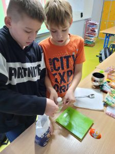 Dwóch uczniów klasy czwartej odmierza cukier przy pomocy łyżeczki do woreczka. Na stole przed nimi stoi waga a przy niej leży opakowanie po kinder jajku.
