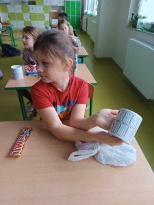Uczennica klasy pierwszej siedzi przy stoliku a w dłoni trzyma kubek otrzymany w prezencie mikołajkowym. Obok na stoliku leży słodki upominek w postaci batona.