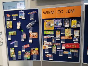 Zdjęcie wystawy zatytułowanej "WIEM CO JEM". Na granatowej dwuskrzydłowej tablicy zamieszczono opakowania po różnych produktach spożywczych z przyczepionymi do nich woreczkami z cukrem.