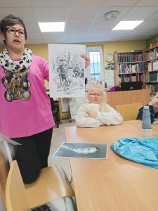 Przy stole za, którym siedzi uczennica klasy pierwszej stoi Pani bibliotekarka. Pani trzyma w dłoni rycinę przedstawiającą scenę z bajki.