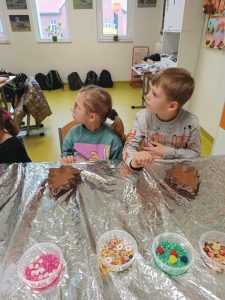 Dwoje uczniów klasy pierwszej siedzi przy stole zakrytym folią aluminiową. Przed dziećmi na stole leżą foremki w kształcie choinek wypełnione czekoladą a dalej kubeczki z kolorowymi ozdobami do czekoladek.