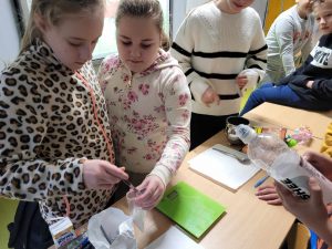 Dwie uczennice odmierzają przy pomocy łyżeczki cukier do strunowego woreczka. Przed nimi stoi na stole waga, a uczeń stojący po drugiej stronie stołu trzyma butelkę po napoju "Oshee".