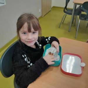 Uczennica oddziału przedszkolnego siedzi przy otwartej śniadaniówce. Dziewczynka wyciąga z papieru swoją kanapkę.