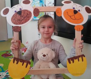 Uczennica oddziału przedszkolnego pozuje do zdjęcia wraz ze swoim misiem. Dziewczynka trzyma drewnianą ramkę ozdobioną w górnych rogach pyszczkami misiów a w dolnych ich łapkami. Misiu dziewczynki opiera się o dolną część ramki.