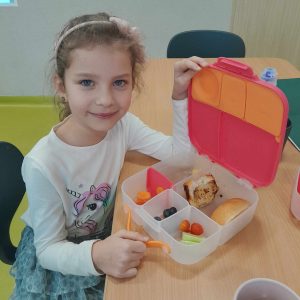 Uczennica oddziału przedszkolnego siedzi przy stole trzymając jedną ręką wieczko śniadaniówki ustawionej przed nią. W śniadaniówce poukładane są owoce i warzywa oraz kawałki kanapek.