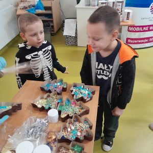 Dwóch uczniów oddziału przedszkolnego stoi przy stoliku na którym leżą gotowe czekolady w kształcie choinek, umieszczone w papierowych foremkach.