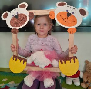 Uczennica oddziału przedszkolnego pozuje do zdjęcia wraz ze swoim misiem. Dziewczynka trzyma drewnianą ramkę ozdobioną w górnych rogach pyszczkami misiów a w dolnych ich łapkami. Misiu dziewczynki opiera się o dolną część ramki.