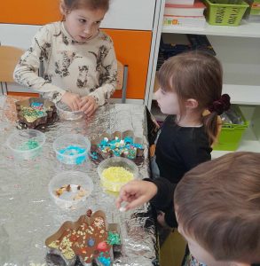 Dwie uczennice oddziału przedszkolnego siedzą za stołem przykrytym aluminiową folią. Na stoliku przed nimi leżą czekolady w foremkach w kształcie choinki oraz pudełka z kolorowymi posypkami do ich ozdobienia.