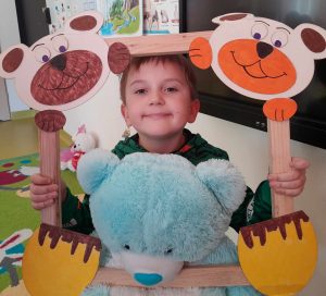 Uczeń oddziału przedszkolnego pozuje do zdjęcia wraz ze swoim misiem. Chłopiec trzyma drewnianą ramkę ozdobioną w górnych rogach pyszczkami misiów a w dolnych ich łapkami. Misiu chłopca opiera się o dolną część ramki.