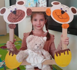 Uczennica oddziału przedszkolnego pozuje do zdjęcia wraz ze swoim misiem. Dziewczynka trzyma drewnianą ramkę ozdobioną w górnych rogach pyszczkami misiów a w dolnych ich łapkami. Misiu dziewczynki opiera się o dolną część ramki.