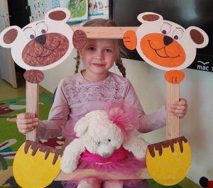 Uczennica oddziału przedszkolnego pozuje do zdjęcia wraz ze swoim misiem. Dziewczynka trzyma drewnianą ramkę ozdobioną w górnych rogach pyszczkami misiów a w dolnych ich łapkami. Misiu dziewczynki opiera się o dolną część ramki.