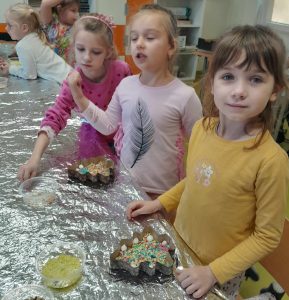 Trzy uczennice oddziału przedszkolnego stoją przy stole przykrytym folią aluminiową. Przed dwiema z nich leżą gotowe czekoladki umieszczone w foremkach w kształcie choinek. Na stoliku leżą przezroczyste pudełka z kolorową posypką.