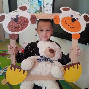 Uczeń oddziału przedszkolnego pozuje do zdjęcia wraz ze swoim misiem. Chłopiec trzyma drewnianą ramkę ozdobioną w górnych rogach pyszczkami misiów a w dolnych ich łapkami. Misiu chłopca opiera się o dolną część ramki.