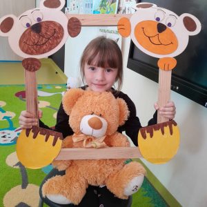 Uczennica oddziału przedszkolnego pozuje do zdjęcia wraz ze swoim misiem. Dziewczynka trzyma drewnianą ramkę ozdobioną w górnych rogach pyszczkami misiów a w dolnych ich łapkami. Misiu dziewczynki opiera się o dolną część ramki.