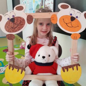 Uczennica oddziału przedszkolnego pozuje do zdjęcia wraz ze swoim misiem. Dziewczynka trzyma drewnianą ramkę ozdobioną w górnych rogach pyszczkami misiów a w dolnych ich łapkami. Misiu dziewczynki opiera się o dolną część ramki.