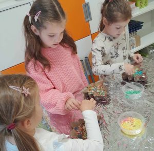 Trzy uczennice oddziału przedszkolnego siedzą za stołem przykrytym aluminiową folią. Na stoliku przed nimi leżą czekolady w foremkach w kształcie choinki oraz pudełka z kolorowymi posypkami do ich ozdobienia.