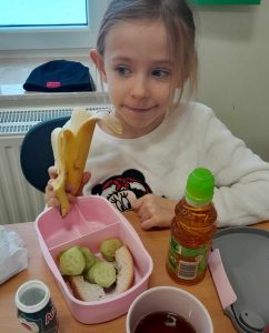 Uczennica oddziału przedszkolnego siedzi przy otwartej śniadaniówce z warzywami. Dziewczynka trzyma w dłoni banana. Obok śniadaniówki stoi soczek.