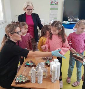 Uczniowie oddziału przedszkolnego wraz z Panią dyrektor stoją przy stoliku za którym siedzi Pani prowadząca warsztaty czekolady. Na stoliku leżą gotowe czekolady w kształcie choinek oraz stoją tubki z ozdobami w sprayu. Pani wyciąga przed siebie dłoń w rękawiczce sięgając po kolejną czekoladkę.