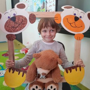 Uczeń oddziału przedszkolnego pozuje do zdjęcia wraz ze swoim misiem. Chłopiec trzyma drewnianą ramkę ozdobioną w górnych rogach pyszczkami misiów a w dolnych ich łapkami. Misiu chłopca opiera się o dolną część ramki.