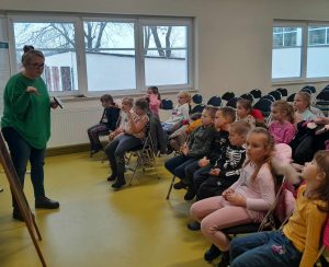 Pani prowadząca warsztaty trzyma w dłoni miernik. Na przeciwko na krzesełkach siedzą uczniowie oddziału przedszkolnego. Zdjęcie wykonane z boku.
