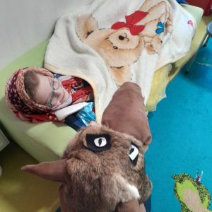 Dziewczynka w stroju babci leży na kanapie. Przykryta jest kocem. Z boku widać głowę wilka - fragment przebrania jednego z chłopców.