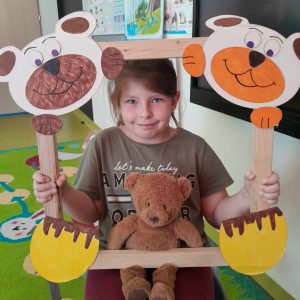 Uczennica oddziału przedszkolnego pozuje do zdjęcia wraz ze swoim misiem. Dziewczynka trzyma drewnianą ramkę ozdobioną w górnych rogach pyszczkami misiów a w dolnych ich łapkami. Misiu dziewczynki opiera się o dolną część ramki.