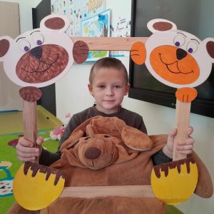 Uczeń oddziału przedszkolnego pozuje do zdjęcia wraz ze swoim misiem. Chłopiec trzyma drewnianą ramkę ozdobioną w górnych rogach pyszczkami misiów a w dolnych ich łapkami. Misiu chłopca opiera się o dolną część ramki.