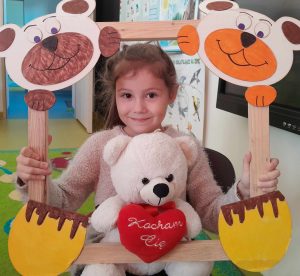 Uczennica oddziału przedszkolnego pozuje do zdjęcia wraz ze swoim misiem. Dziewczynka trzyma drewnianą ramkę ozdobioną w górnych rogach pyszczkami misiów a w dolnych ich łapkami. Misiu dziewczynki opiera się o dolną część ramki.