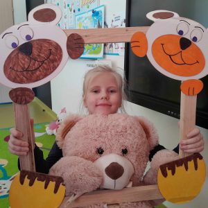 Uczennica oddziału przedszkolnego pozuje do zdjęcia wraz ze swoim misiem. Dziewczynka trzyma drewnianą ramkę ozdobioną w górnych rogach pyszczkami misiów a w dolnych ich łapkami. Misiu dziewczynki opiera się o dolną część ramki.