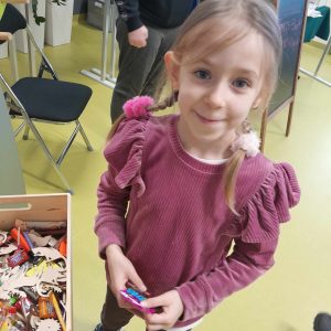 Uczennica oddziału przedszkolnego stoi obok drewnianego pudełka wypełnionego drobnymi upominkami dla dzieci. Dziewczynka trzyma w dłoniach wybraną przez siebie nagrodę.
