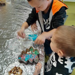 Dwóch uczniów oddziału przedszkolnego pracuje obok siebie nad swoimi czekoladami. Chłopcy siedzą przy stole przykrytym folią aluminiową a przed nimi leży ozdabiana czekolada w foremce w kształcie choinki. Chłopcy wyciągają nad nią dłonie trzymając w nich kolorowe posypki.