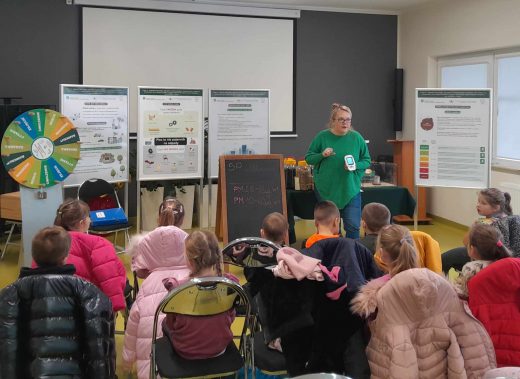 Pani prowadząca warsztaty stoi trzymając w dłoniach miernik. Obok niej ustawione są na sztalugach plakaty informacyjne. Na przeciwko siedzą uczniowie.