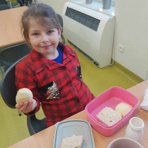 Uczennica oddziału przedszkolnego siedzi przy otwartej śniadaniówce z warzywami. Dziewczynka trzyma w dłoni obrane jabłko.