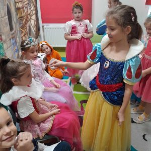 Zdjęcie wykonane podczas przedstawienia. Dziewczynka w stroju wróżki dotyka różdżką głowy siedzącej na przeciwko uczennicy przebranej za księżniczkę. Obok niej siedzi dziewczynka z koroną na głowie.