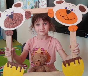 Uczennica oddziału przedszkolnego pozuje do zdjęcia wraz ze swoim misiem. Dziewczynka trzyma drewnianą ramkę ozdobioną w górnych rogach pyszczkami misiów a w dolnych ich łapkami. Misiu dziewczynki opiera się o dolną część ramki.