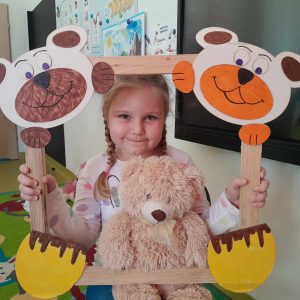 Uczennica oddziału przedszkolnego pozuje do zdjęcia wraz ze swoim misiem. Dziewczynka trzyma drewnianą ramkę ozdobioną w górnych rogach pyszczkami misiów a w dolnych ich łapkami. Misiu dziewczynki opiera się o dolną część ramki.