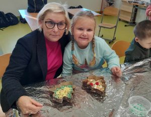 Pani dyrektor wraz z uczennicą oddziału przedszkolnego pozują do wspólnego zdjęcia. Siedzą obok siebie przy stole przykrytym folia aluminiową a przed nimi na stole leżą gotowe czekolady w foremkach w kształcie choinek.