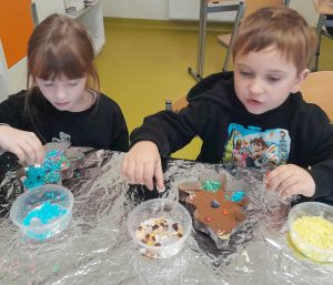 Dwoje uczniów oddziału przedszkolnego siedzi za stołem przykrytym aluminiową folią. Na stoliku przed nimi leżą czekolady w foremkach w kształcie choinki oraz pudełka z kolorowymi posypkami do ich ozdobienia. Chłopiec sięga dłonią do jednego z pojemników, a dziewczynka posypuje swoją czekoladą niebieską posypką.