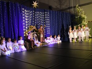 Zdjęcie sceny na której widać szopkę wraz z Maryją i stojącym Józefem. Otaczają ich po bokach dzieci ubrane na biało trzymające białe lampiony. Po prawej stronie stoi pięć dziewczynek w strojach aniołków za złożonymi dłoniami.