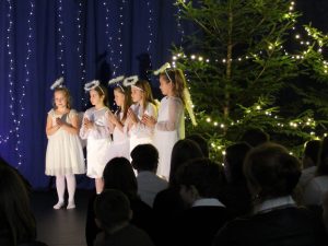 Pięć uczennic w strojach aniołków stoi w rzędzie. Dziewczynki mają złożone dłonie. Za nimi widać choinkę z jasnymi lampkami.