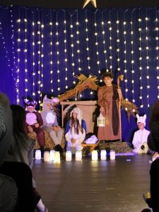 Zdjęcie szopki ustawionej po środku sceny. W szopce siedzi Maryja, obok stoi Józef trzymający pastorał i lampion. Po lewej stronie na snopku siana siedzi dwoje uczniów w strojach zwierzątek. Przed szopką ustawione są w rzędzie lampiony.